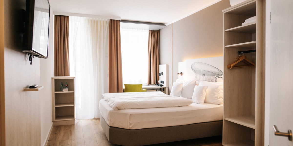 arrierearmes Doppelzimmer im Kongresshotel Potsdam. Der Blick von der Eingangstür in das Zimmer hinein. Ein Doppelbett, ein Fernseher an der Wand, offene Schränke und ein Schreibtisch direkt vor dem Fenster.