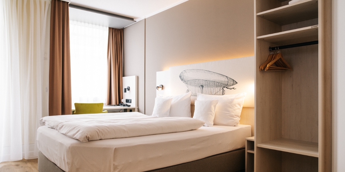 Barrierearmes Doppelzimmer im Kongresshotel Potsdam. Links ist der Schreibtisch vor dem Fenster, in der Mitte das Doppelbett und rechts ein offener Schrank mit Kleiderbügeln.