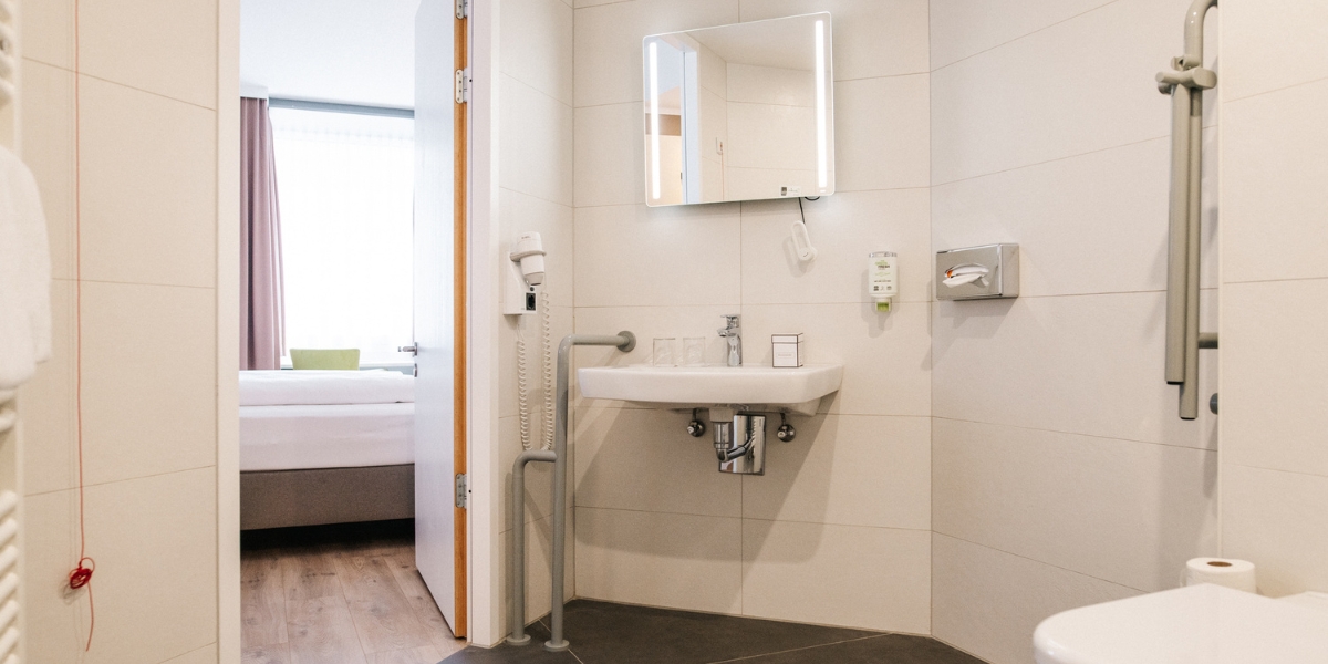 Barrierearmes Badezimmer des Doppelzimmers im Kongresshotel Potsdam. Helle Wandfliesen und dunkle Bodenfliesen. Links ist die Tür zum Schlafzimmer, in der Mitte das Waschbecken mit Seitenhalterung und höhenverstellbarem Spiegel.
