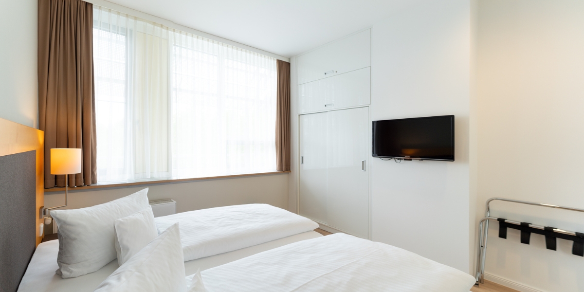arrierearmes Studio im Kongresshotel Potsdam. Blick von der Seite des Bettes auf das große Fenster, dem eingebauten Kleiderschrank und den Fernseher an der Wand.
