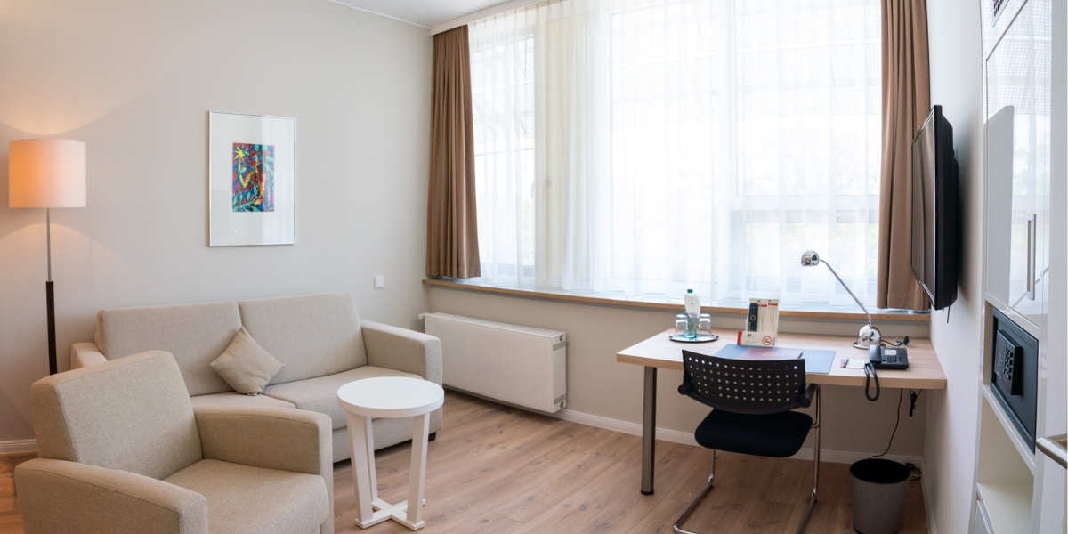 Barrierearmes Studio im Kongresshotel Potsdam. Blick auf den Wohnbereich mit einer Couch, einem Sessel und einem Schreibtisch direkt vor dem Fenster.