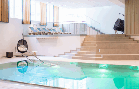 Kongresshotel-Potsdam-Wellbeing-Area-Pool eleuchteter Indoor-Pool mit grün schimmerndem Wasser, links Sitzschaukel und im Hintergrund Treppe zum Ruheraum mit Liegen und Tageslichteinfall