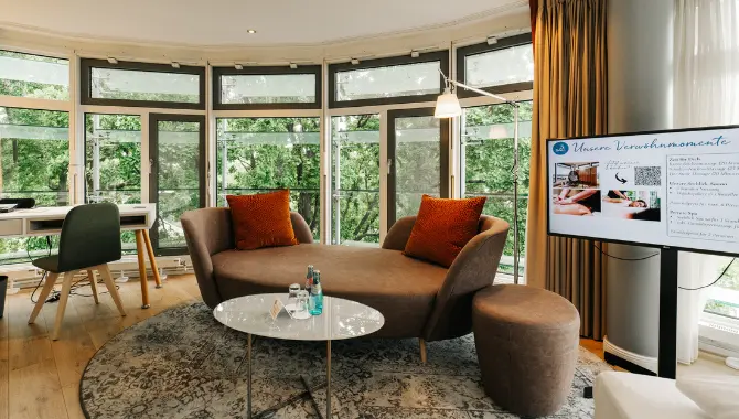 Heller Wohnbereich mit großer Fensterfront, Sofa mit Kissen, kleinem Tisch mit Wasserflaschen sowie Schreibtisch und Bildschirm mit Wellnessangeboten.