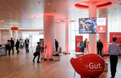 Teilnehmende bewegen sich durch den lichtdurchfluteten Veranstaltungsfoyer mit roter Beleuchtung und Sparkassen-Branding.