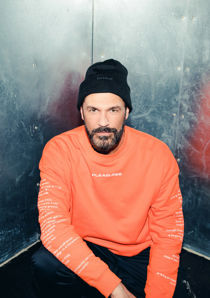 Mann mit Mütze und orangem Sweatshirt sitzt vor dunkler Metallwand.