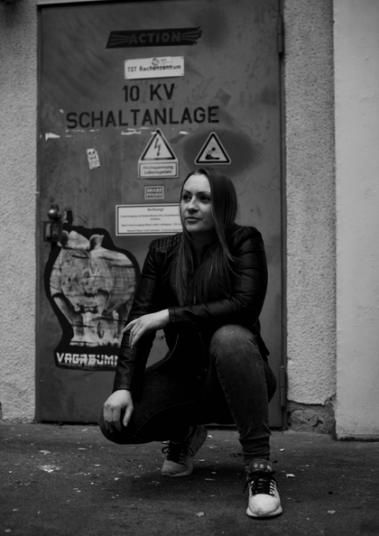 Frau in Lederjacke sitzt vor einer Tür mit Warnschildern einer Schaltanlage.