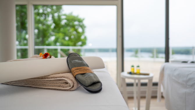 Kongresshotel-Potsdam-Seeblick-Sauna (2) Massageliege mit Handtüchern und Hausschuhen in der Seeblick Sauna. Große Panoramafenster eröffnen einen weiten Blick auf den See.