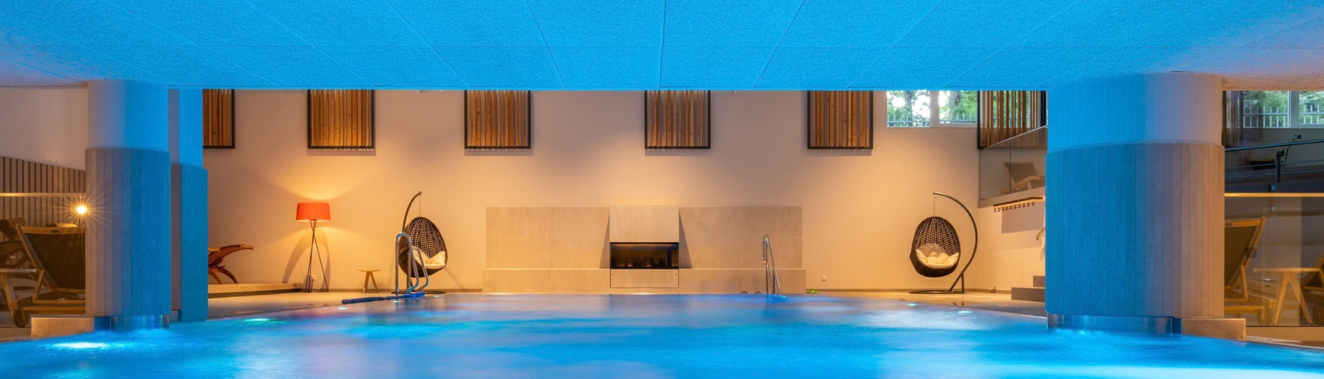 Großer Innenpool in der Wellbeing Area des Kongresshotel Potsdam. Das Wasser ist in blaues Licht getaucht, während warme Beleuchtung, Loungemöbel und ein Kamin für eine entspannte Atmosphäre sorgen. Große Fenster geben den Blick ins Grüne frei.