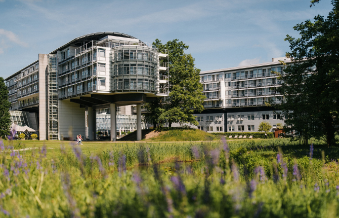 Außenansicht des Kongresshotels Potsdam mit markanter Architektur, umgeben von weitläufigen Grünflächen und blühenden Wiesen in sommerlicher Atmosphäre.