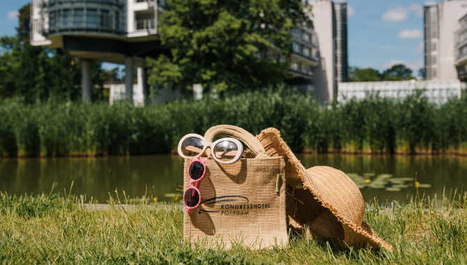 Strandtasche mit Sonnenbrillen und Sonnenhut auf einer Wiese am Wasser vor dem Kongresshotel Potsdam. Die Szene vermittelt entspannte Urlaubstage in naturnaher Umgebung.